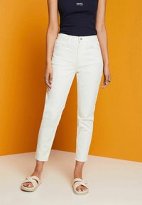 Jeans de denim blanco con corte ajustado y largo hasta el tobillo, combinados con una camiseta sin mangas azul marino y sandalias de deslizar beige sobre un fondo naranja.