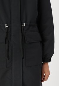 Manteau long noir avec taille à cordon, poignets élastiqués et deux poches avant. Fabriqué en tissu mat et lisse avec une texture subtile.