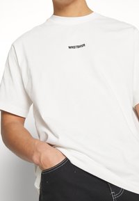 T-shirt en coton blanc à manches courtes, avec un petit logo brodé noir "WRSTBHR" près du col. Design simple et classique.