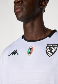 Hvit fotballtrøye med svart krage og detaljer; har Kappa-logoen og Spezia SC-emblemet, samt detaljer med italiensk ligamerke.
