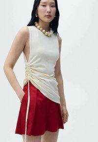Cream sleeveless top with gathered side detail, paired with red pleated shorts. A bead necklace and gold earrings complete the outfit.