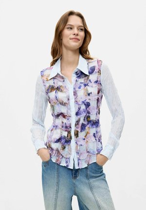 Young woman with hands in pockets wearing a striped shirt with layered purple floral patterns and light blue jeans against a white background.