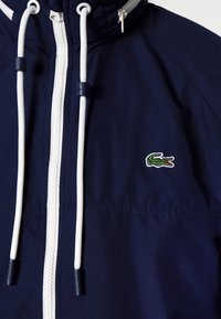 Veste marine avec fermeture éclair blanche et cordons de serrage, arborant un logo crocodile vert. Tissu lisse avec une capuche structurée et des éléments ajustables.