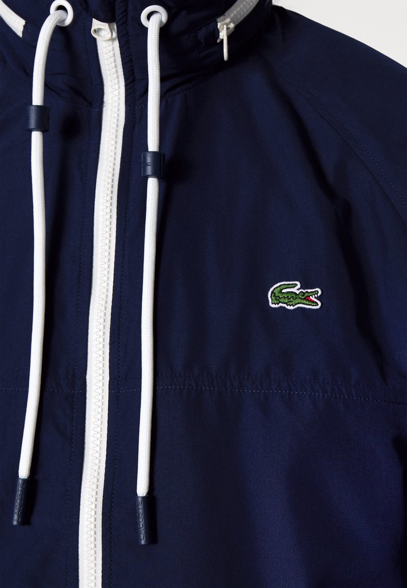 Giacca navy con zip bianca e lacci, caratterizzata da un logo di coccodrillo verde. Tessuto morbido con cappuccio strutturato e elementi regolabili.