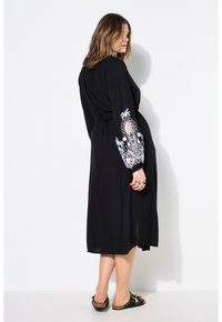Schwarzes Kleid mit lockerer Passform, langen Ärmeln, die mit weißer Floralstickerei versehen sind, und einem Bindegürtel in der Taille. Getragen mit flachen, schwarzen Sandalen.