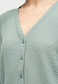 Πράσινο ανοιχτόχρωμο υφασμάτινο τοπ με κουμπιά και υφή, με λαιμόκοψη σε σχήμα V στο πάνω μέρος του θώρακα και του λαιμού ενός ατόμου.