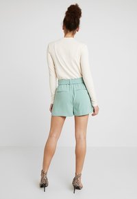 Svetle zelené kraťasy s měkkou strukturou, elastickým pasem a dvěma zadními kapsami, doplněné krémovým topem s dlouhým rukávem a vzorovanými podpatky.