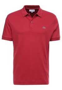 Rood katoenen poloshirt met korte mouwen, twee knopen aan de sluiting en een klein groen Lacoste-logo op de linkerborst. Soepele textuur.
