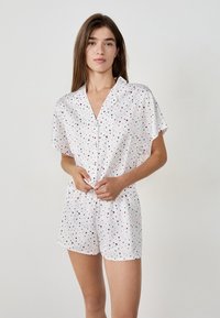 Jonge vrouw in witte shorts en een button-up top met kleine roze en zwarte hartjespatronen op een effen achtergrond.