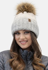 Gorro de punto gris y crema con un gran pompón de piel sintética en la parte superior y una etiqueta color marrón. Presenta una textura acanalada para mayor interés.