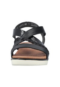 Svart sandal med korsremmar och flätade texturer. Materialen inkluderar syntetisk läder och en ljusfärgad sula. Flat och öppen tå.