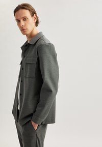 Chaqueta gris de botones con cuello, dos bolsillos en el pecho y una tela suave y texturizada. Llevada sobre una camisa blanca, combinada con pantalones a juego.