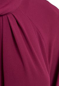 Tessuto color prugna con una finitura morbida e strutturata. Presenta pieghe lungo un lato, in evidenza una caduta fluida e cuciture su misura.