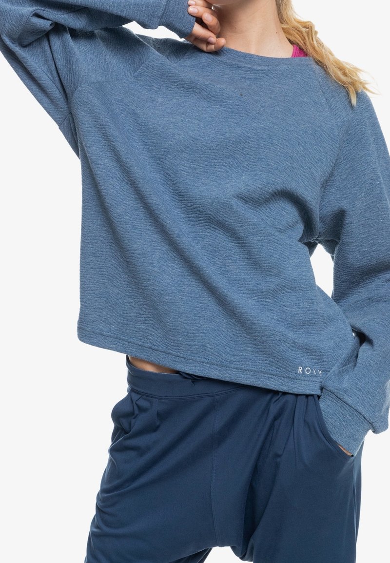 Blue, textured sweatshirt with a loose fit and dropped shoulders. Features Roxy logo on the hem. Paired with dark blue, relaxed-fit pants.