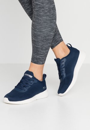 Osoba nosząca ciemnoszare legginsy i lekkie granatowe buty sportowe z białymi podeszwami, stojąca na jasnoszarej podłodze na jednolitym tle.