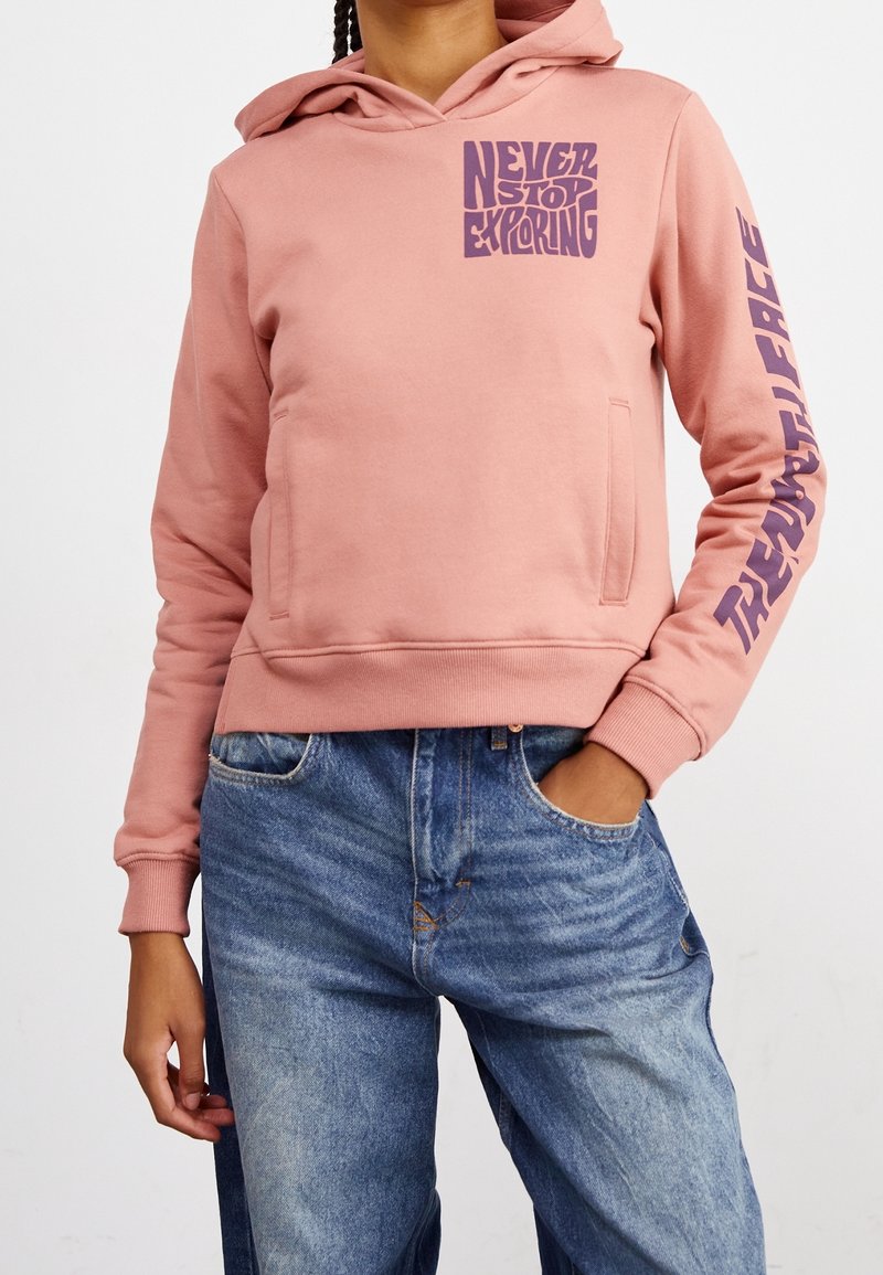 Personne portant un sweat à capuche rose avec un texte violet « Never Stop Exploring » sur la poitrine et « Travel Wherever » sur la manche gauche, associé à un jean bleu.