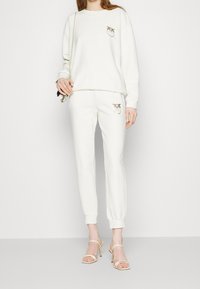 Woman wearing white sweatshirt and matching joggers with gold bird logos, holding small metallic handbag, and wearing white strappy heels.