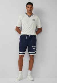 Witte v-hals shirt met marineblauwe rand, gecombineerd met marineblauwe drawstring shorts met witte accenten en een palmboomgrafiek, staand op een neutrale achtergrond.