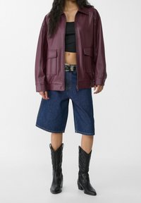 Burgundy läderjacka med dragkedja och två framfickor, kombinerad med vida denimshorts och svarta cowboystövlar.