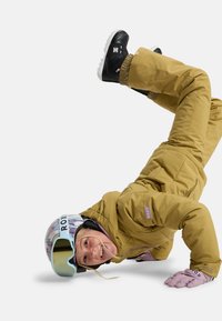 Gelbe isolierte Skiausrüstung mit Kapuze, kombiniert mit schwarzen Schneeschuhen. Ein mehrfarbiger Helm und pinke Handschuhe vervollständigen den Look.