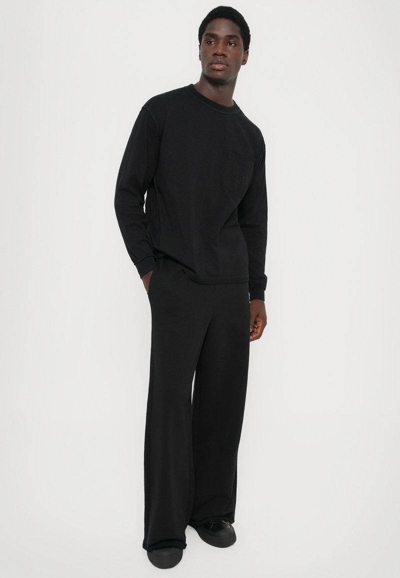 Chemise noire à manches longues avec une poche poitrine, accompagnée de pantalons larges noirs. Le tissu semble doux et lisse, avec une coupe décontractée.