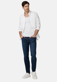 Biała koszula z guzikami noszona na białym topie na ramiączkach, w zestawieniu z ciemnoniebieskimi dżinsami i białymi sneakersami. Stylizacja ma swobodny krój.