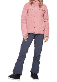 Rosa Cordjacke mit Knopfverschluss, Brusttaschen und hohem Kragen, kombiniert mit grauen Skihosen und schwarzen Stiefeln mit weißen Sohlen.