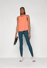 Κοραλλί αθλητικό μπλουζάκι με στρογγυλή λαιμόκοψη και χαλαρή εφαρμογή, συνδυασμένο με τυρκουάζ κολάν. Κρατώντας μια μαύρη φιάλη νερού, φ wearing ελαφριά μωβ παπούτσια.