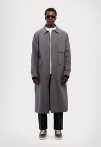 Grey long coat with buttons, featuring a front pocket and a tailored fit. Worn over a white shirt and paired with dark trousers and sneakers.