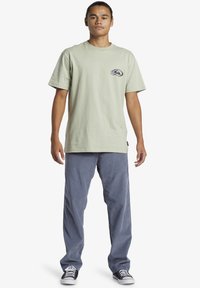 Light green cotton t-shirt with a crew neck, featuring a small floral graphic on the left chest. Paired with blue corduroy pants and black sneakers.