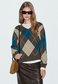 Brun och teal argyle-mönstrad v-ringad tröja med krämfärgad detaljer, bärs över en vit topp, ihop med en brun läderkjol och väska.