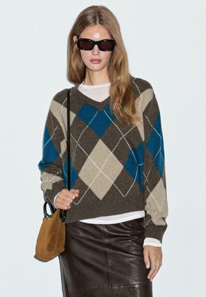 Bruine en teal argyle-gepatroneerde v-hals trui met crèmekleurige details, gedragen over een witte top, gecombineerd met een bruine leren rok en tas.