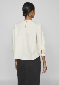 Blouse en coton couleur crème avec des manches longues bouffantes, détail de boutons décoratif au dos et dentelle aux ouvertures des manches.
