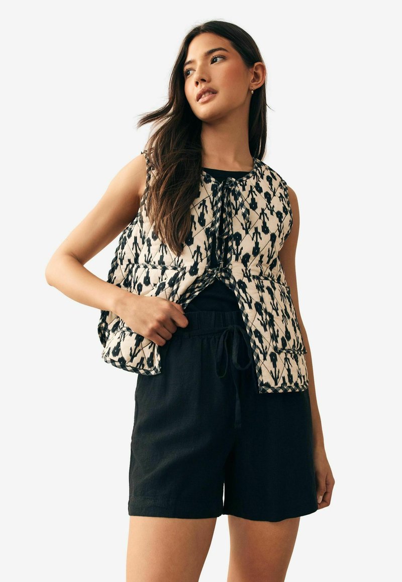 Chaleco acolchado en marfil con un patrón geométrico negro; presenta un cuello redondo y un frente abierto. Combinado con pantalones cortos negros con cordón.