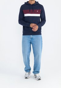 Man som bär en marinblå hoodie med vinröda och vita ränder, ljusblå jeans och silverfärgade löparskor, står med händerna sammanflätade.