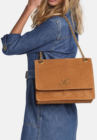 Blonde woman wearing a blue denim dress carries a large tan suede handbag with a gold chain strap and gold logo clasp.