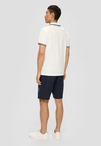 Chemise polo blanche avec des accents de rayures noires au niveau du col ; accompagnée de shorts bleu foncé avec un motif floral ; baskets blanches.