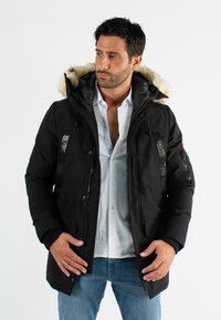 Parka noire avec capuche en fourrure, fermeture éclair à l'avant, plusieurs poches et tissu texturé. Présente des accents de marque et des poignets côtelés.