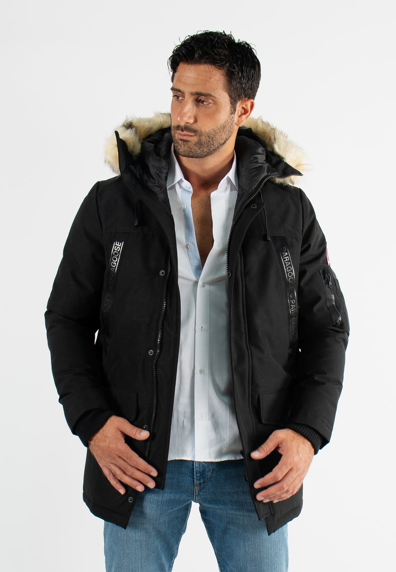 Parka noire avec capuche en fourrure, fermeture éclair à l'avant, plusieurs poches et tissu texturé. Présente des accents de marque et des poignets côtelés.