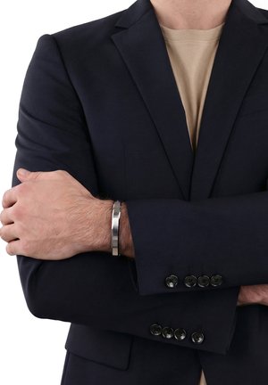 Man met een donkerblauwe blazer en beige overhemd met gekruiste armen, die een zilverkleurige metalen armband aan zijn linker pols toont.