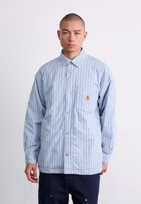 Chemise bleu clair à manches longues, à rayures verticales fines, avec une poche poitrine arborant un logo et une coupe décontractée.