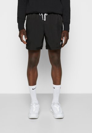 Uomo con pantaloncini neri Nike con coulisse bianca, calzini Nike bianchi e scarpe da ginnastica bianche, in piedi su uno sfondo semplice.