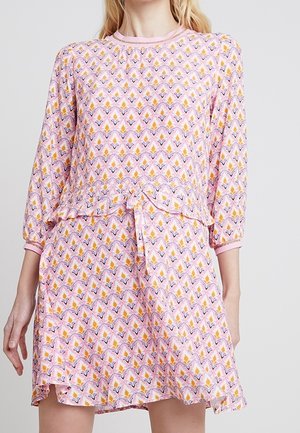Kobieta ubrana w różową sukienkę w wzory, z rękawami do łokcia i rozkloszowanym dołem, ozdobioną małą falbanką w talii.
