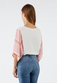 Kortærmet top med hvid krop og røde mønstrede ærmer med zigzag- og rutemønstre, parret med blå denim jeans.