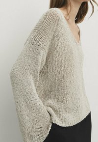 Beige knitted sweater with a deep V-neck, wide sleeves, and a loose fit; textured yarn and visible stitching details, paired with a black bottom.