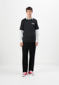 Young man wearing black "Empire" t-shirt over white striped shirt, black pants, and red sneakers, standing against a plain white background.