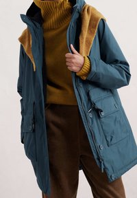 Tyrkysový voděodolný kabát s kapucí, se zipem a knoflíky, přetáhnutý přes hořčicový svetr. Viditelné texturované hnědé kalhoty z kordu.