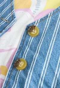 Denim odjevni predmet s vertikalnim bijelim prugama, s dva okrugla, prozirna žuta gumba. Mekani, šareni materijal ispod.