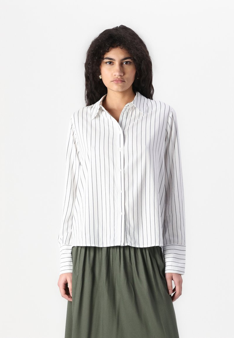 Chemise blanche à boutons avec des rayures verticales navy, col, manches longues, et un ourlet légèrement court, associée à une jupe verte.
