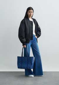 Sinine kandekott mustrihise kujundusega, mida kannab inimene, kes on seljas must bomber-jakk, valge särk ja laia säärega sinised teksad.